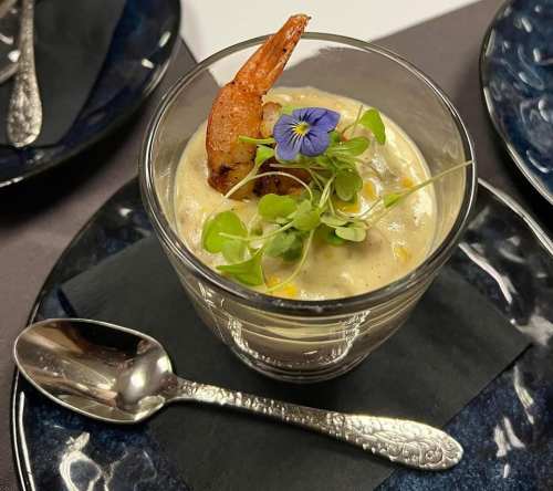 A gourmet dish in a glass, featuring shrimp, edible flowers, and microgreens, served on a dark plate with a spoon.