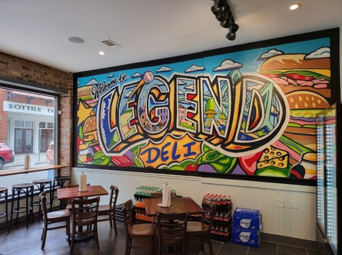 Colorful mural reading "Welcome to Legend Deli" on a restaurant wall, with tables and drinks in the foreground.