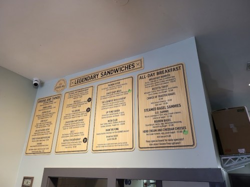 Menu board featuring various sandwich options and all-day breakfast items, displayed on a wooden background.