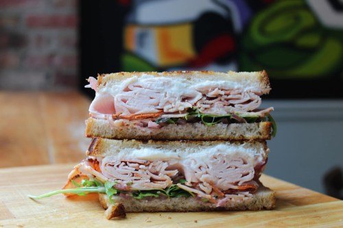 A stacked sandwich with layers of turkey, greens, and sliced vegetables on toasted bread, set on a wooden surface.