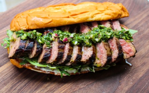 A grilled steak sandwich with sliced meat, topped with green sauce and served on a toasted bun with greens.