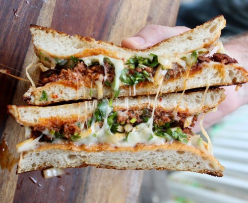A hand holding a stacked sandwich filled with meat, cheese, and greens, with melted cheese stretching between the layers.