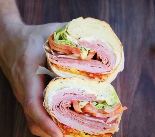 A hand holding a large sandwich filled with layers of ham, lettuce, tomato, and condiments on a wooden surface.