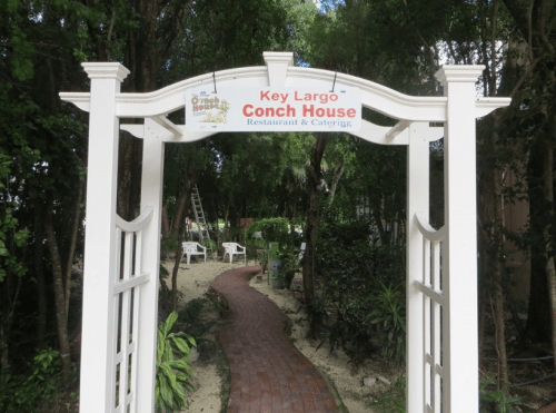 A white archway with a sign reading "Key Largo Conch House Restaurant & Catering" surrounded by greenery.