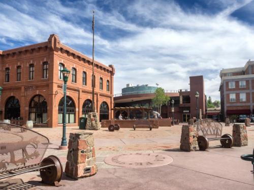 A sunny plaza featuring brick buildings, benches, and stone sculptures under a blue sky with scattered clouds.