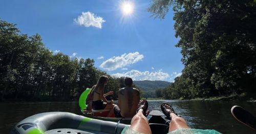 Two people relax on a raft in a river, surrounded by trees and under a bright sun with a clear blue sky.