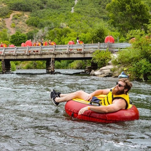 A person in a yellow life jacket relaxes on a red inner tube in a river, with a bridge and other tubers in the background.