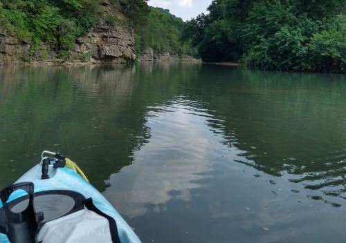 A serene river scene viewed from a kayak, surrounded by lush greenery and rocky cliffs under a cloudy sky.