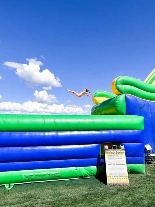 A person jumps off a colorful inflatable slide against a blue sky with fluffy clouds.