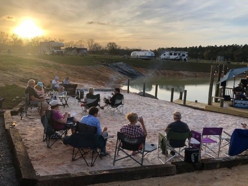 A group of people sitting in chairs by a sandy area near a lake, enjoying a sunset and live music. RVs are in the background.