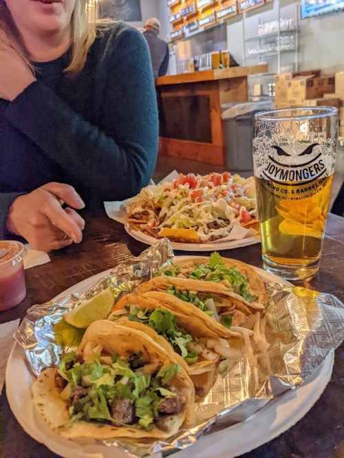 A table with tacos topped with cilantro and lime, a plate of nachos, and a glass of beer in a lively restaurant setting.