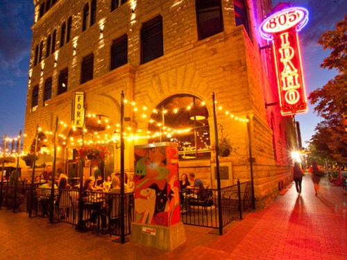 A lively restaurant with outdoor seating, string lights, and a colorful mural, set against a historic building at dusk.