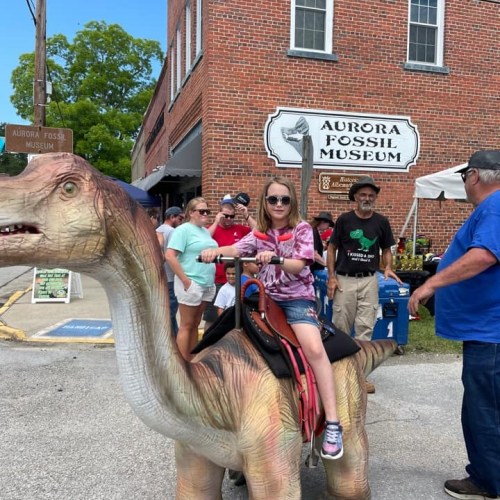 A girl in a tie-dye shirt rides a dinosaur statue outside the Aurora Fossil Museum, with people gathered around.