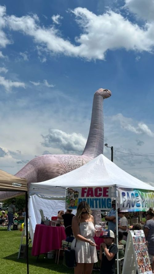 A large inflatable dinosaur towers over a festival scene with tents and people enjoying activities below.