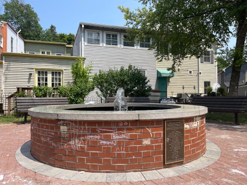 A circular brick fountain with a small water feature, surrounded by benches and houses in a sunny park setting.