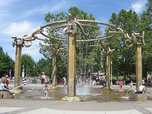 A decorative fountain with water jets, surrounded by trees and people enjoying the park on a sunny day.