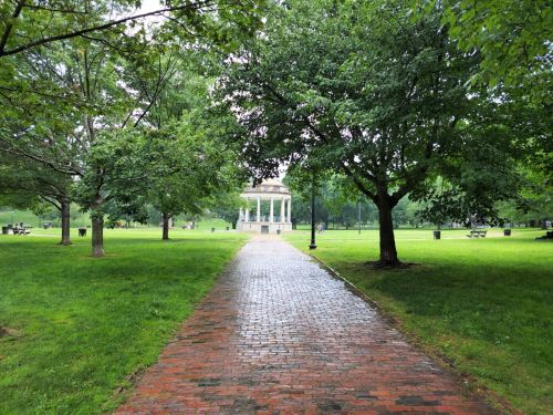 A brick path leads through a lush green park, with trees on either side and a gazebo in the distance.