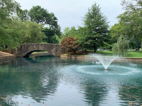 A serene park scene featuring a stone bridge over a pond with a fountain and lush green trees surrounding the area.