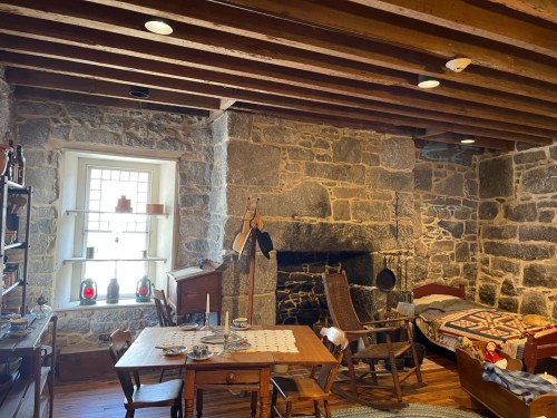 Cozy room with stone walls, wooden beams, a dining table, rocking chair, and a bed, featuring vintage decor.