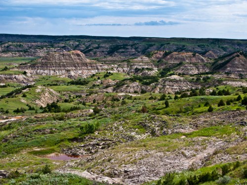 A panoramic view of rolling hills and rugged terrain under a cloudy sky, with patches of greenery and a small pond.