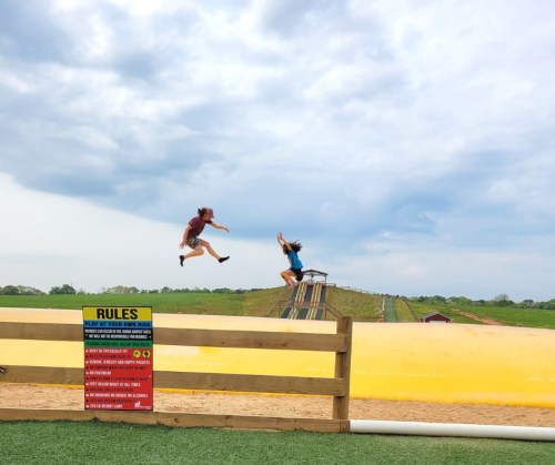 Two people jump joyfully in the air near a large inflatable slide, with a sign listing rules in the foreground.