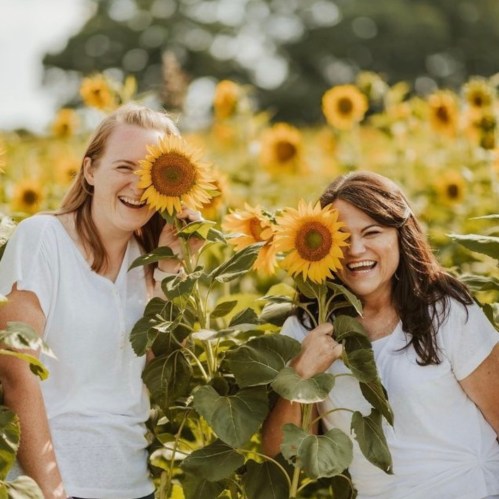 Two women smiling and playfully hiding behind large sunflowers in a vibrant sunflower field.