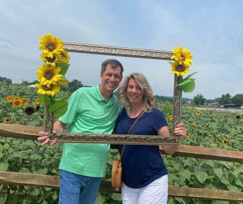 A couple poses with a decorative frame adorned with sunflowers in a sunflower field.