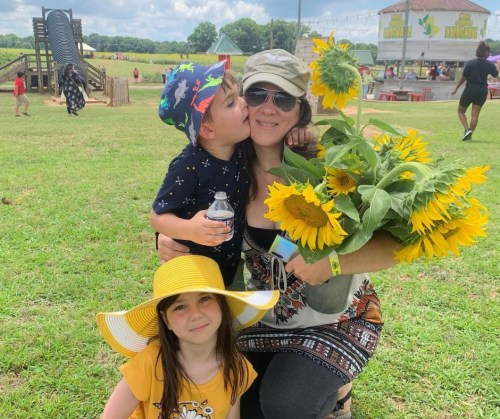 A woman holds a bouquet of sunflowers, smiling with a child kissing her cheek and another child sitting beside her.