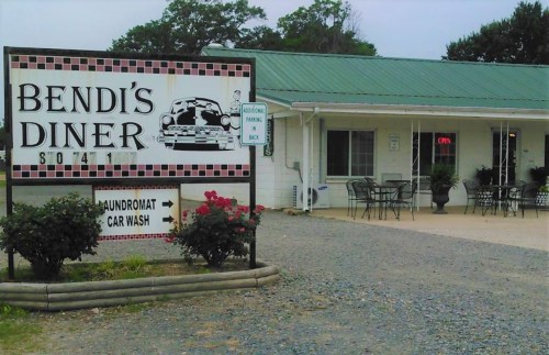 A roadside diner with a sign reading "Bendi's Diner," featuring outdoor seating and a laundromat and car wash nearby.