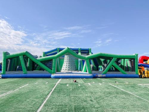 A large green and blue inflatable obstacle course set up on a grassy field under a clear blue sky.