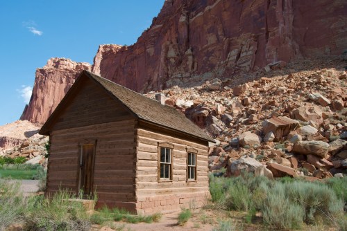 A small wooden cabin stands in front of a rocky cliff, surrounded by sparse vegetation under a clear blue sky.