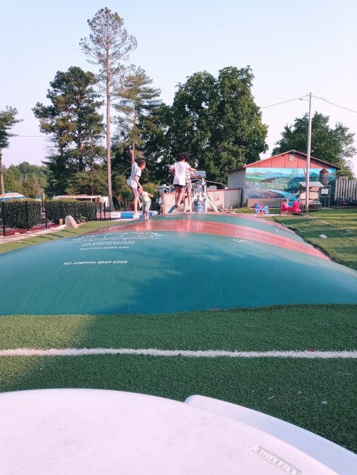 Two children play on a large inflatable bounce pad in a grassy area, surrounded by trees and a colorful backdrop.