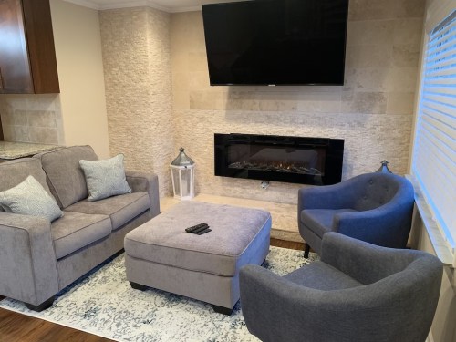 Cozy living room with a gray sofa, armchair, ottoman, and a modern fireplace beneath a wall-mounted TV.