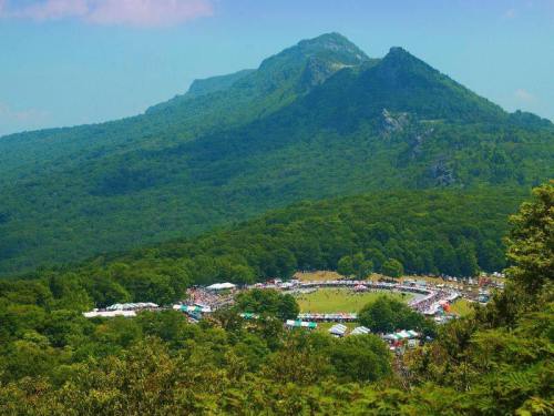 A panoramic view of a lush green mountain landscape with a gathering or festival in a valley below.