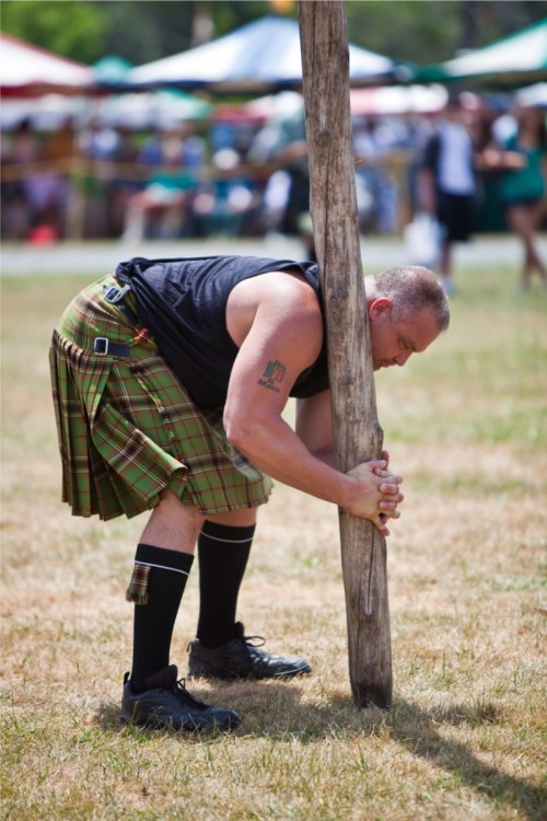 A man in a kilt prepares to lift a wooden pole at an outdoor festival, surrounded by tents and spectators.
