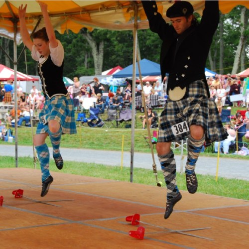 Two dancers in traditional Scottish attire perform on stage, showcasing energetic movements under a colorful tent.