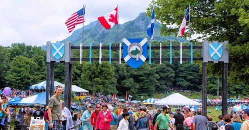 A festival entrance with flags and a mountain backdrop, bustling with people enjoying the event.