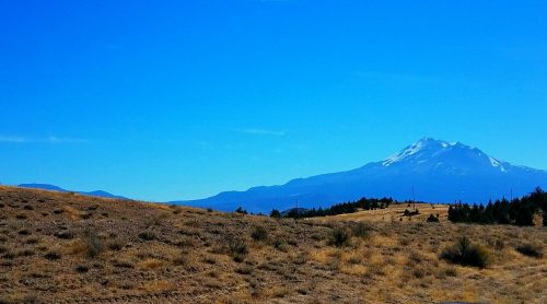 A clear blue sky over a mountainous landscape with a snow-capped peak and dry, grassy terrain in the foreground.