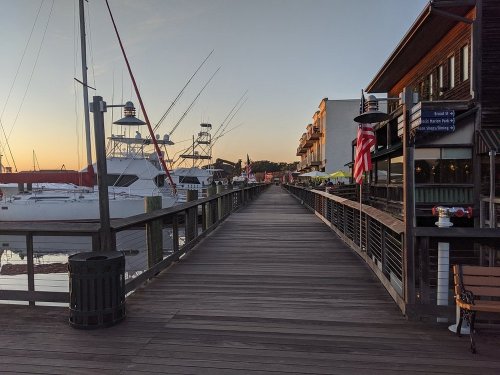 A wooden pier lined with boats and buildings, bathed in warm sunset light. Flags and outdoor seating are visible.
