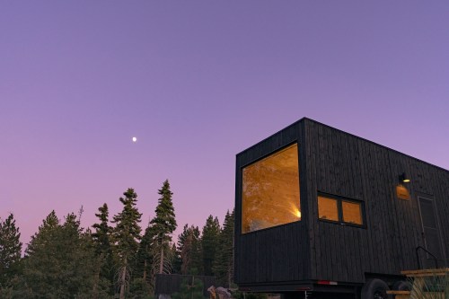 A modern black tiny house surrounded by trees under a twilight sky with a bright moon.