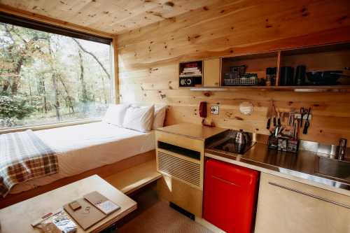 Cozy wooden cabin interior with a bed, kitchenette, and large window overlooking a forest.