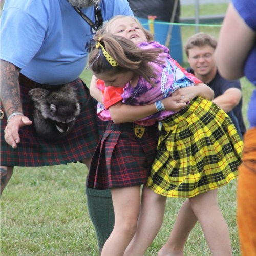 A man in a kilt holds a girl in a colorful skirt while another girl playfully leans against him at an outdoor event.