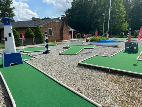 A mini-golf course featuring colorful obstacles like lighthouses and boats, surrounded by green turf and trees.