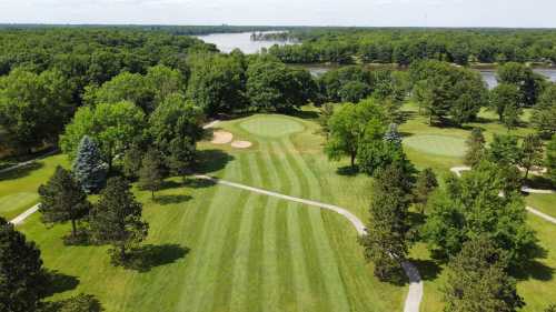 Aerial view of a lush green golf course with sand traps, surrounded by trees and a river in the background.