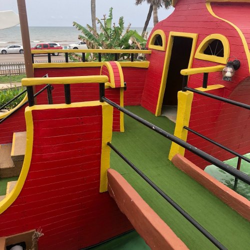 Colorful play structure resembling a pirate ship, with a green walkway and ocean view in the background.