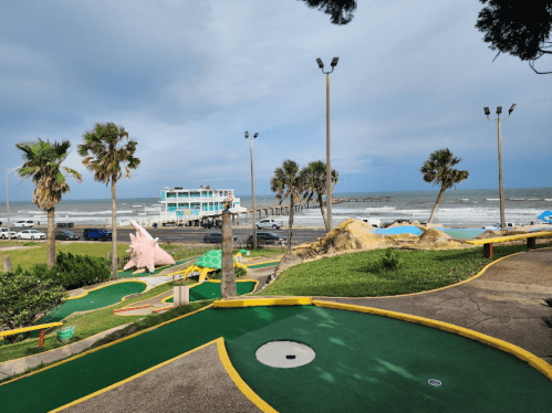 A mini-golf course with palm trees, overlooking the ocean and a pier under a cloudy sky.