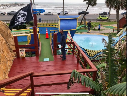 A man stands on a wooden deck overlooking a colorful mini-golf course near the beach, with palm trees and a pirate flag.