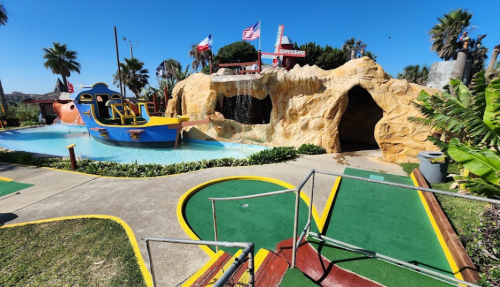 Colorful mini-golf course featuring a pirate ship, waterfall, and rocky landscape under a clear blue sky.