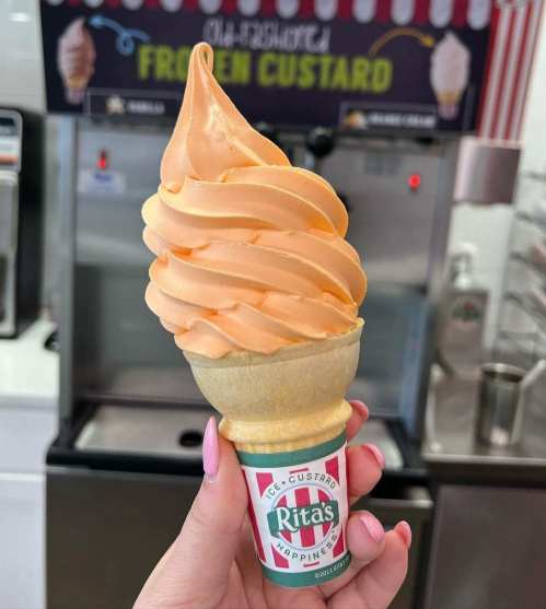 A hand holding a cone of orange frozen custard from Rita's, with a colorful sign in the background.