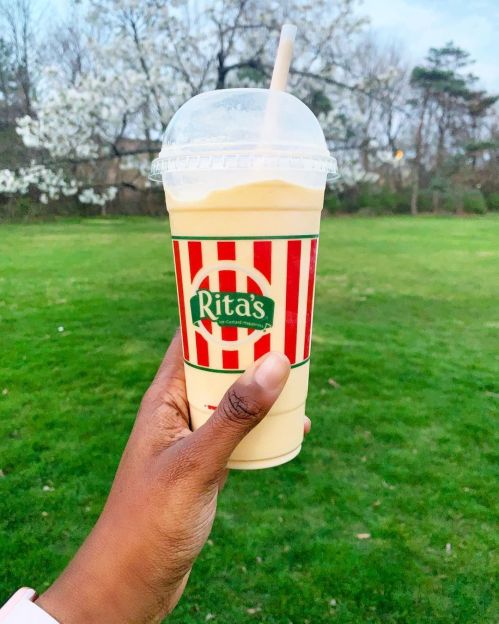 A hand holding a Rita's frozen treat cup with a yellow drink, set against a green park background.
